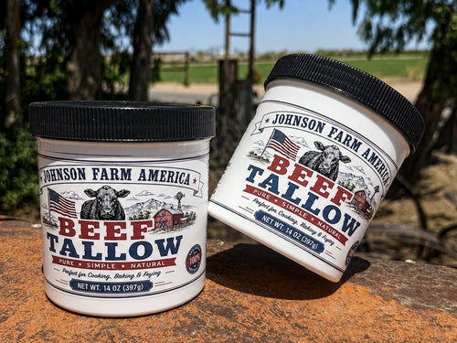 Two white plastic tubs of Johnson Farm America Beef Tallow lard, one standing upright and the other tilted, set on a rustic orange-brown wooden surface with an outdoor farm background.